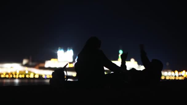 Unga par sitter på stranden och tittar på natten stadsutsikt — Stockvideo
