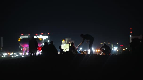 Молодые люди устраивают вечеринку на пляже ночью на фоне светящихся небоскребов - курящий кальян — стоковое видео