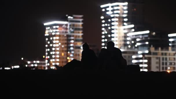Giovane uomo e donna seduti sulla spiaggia di notte sullo sfondo di edifici moderni incandescenti e parlando tra loro — Video Stock