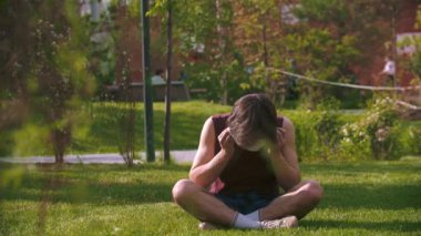 Genç bir adam çimlerin üzerinde oturuyor ve sağlık maskesi takıyor.