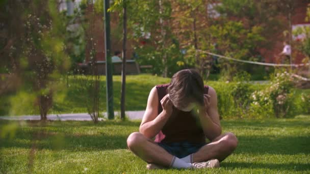 Ein junger Mann sitzt auf dem Rasen und setzt sich die medizinische Maske auf — Stockvideo