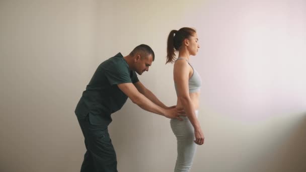 Žena podstupující léčbu osteopatem - stojí na podlaze a ohýbá se, zatímco ji doktor drží za boky