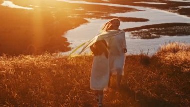 Genç anne beyaz elbiseli kızıyla tepede koşuyor ve gün batımında uçurtmayla oynuyor.