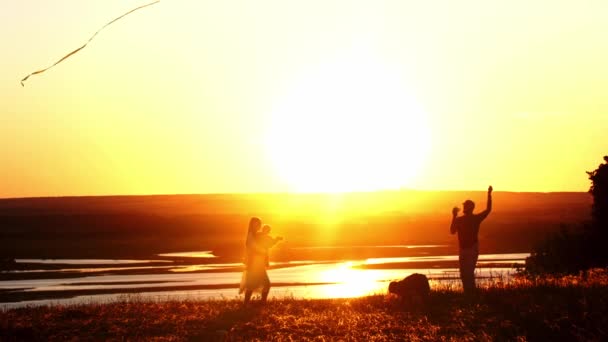 Νεαρή οικογένεια παίζει με ένα χαρταετό στο λόφο, ενώ το φωτεινό πορτοκαλί ηλιοβασίλεμα — Αρχείο Βίντεο