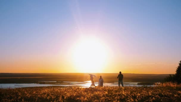 Família jovem em pé na colina em um pôr do sol cedo - uma menina brincando com o papagaio — Vídeo de Stock