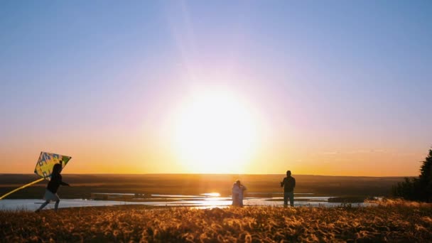 Familia joven de pie en la colina en una puesta de sol temprana - una niña jugando con la cometa y corriendo por el campo — Vídeo de stock