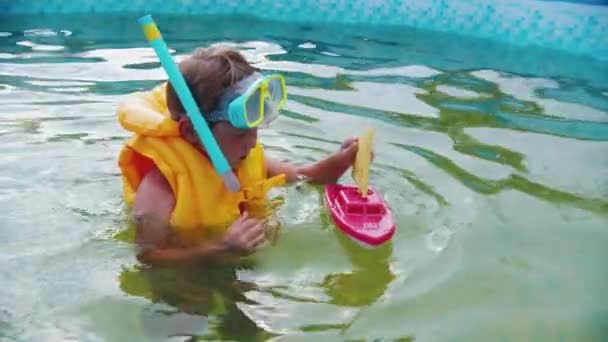 Маленький мальчик в подводной маске играет с игрушечной лодкой в надувном бассейне — стоковое видео