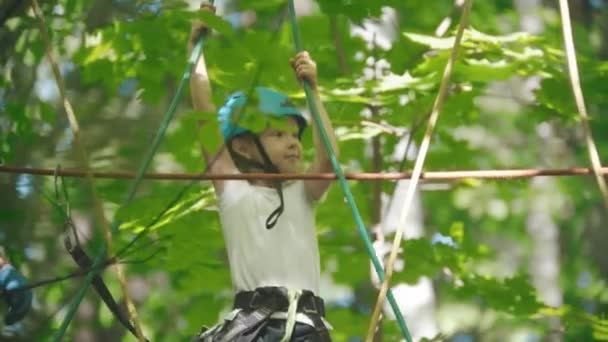 Een klein meisje dat over de brug loopt en aan de touwen vasthoudt. Een amusementsattractie in het groene bos. — Stockvideo