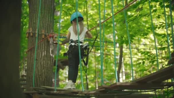 Een klein meisje dat over de touwbrug loopt en aan de touwen vasthoudt. Een amusementsattractie in het groene bos. — Stockvideo