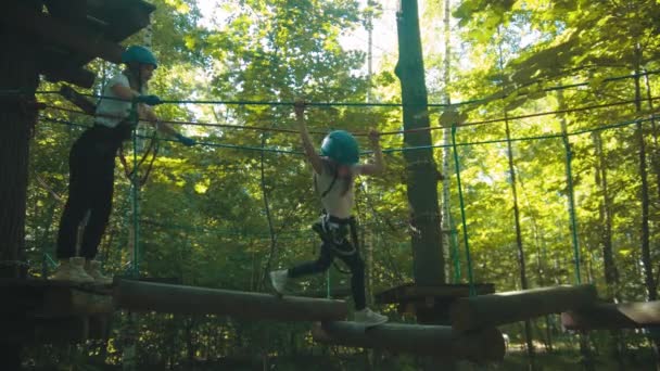 Jovem e sua filha em uma atração no parque a menina andando sobre os troncos pendurados em cordas — Vídeo de Stock