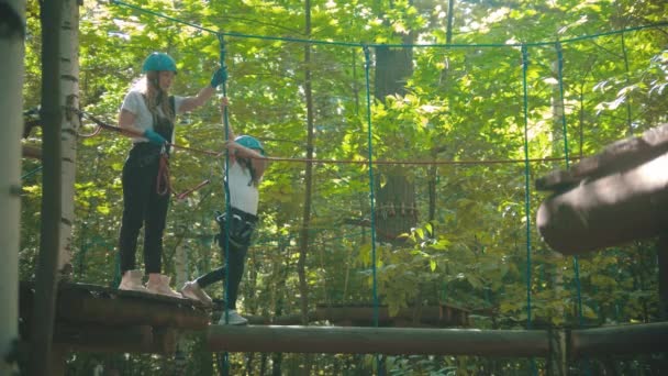Jonge vrouw en haar dochter op een attractie in het bos - het meisje lopen op de logs opknoping op touwen — Stockvideo