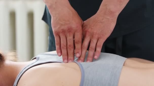Молодая женщина лежит на диване в клинике и массажист давит пальцами на ее позвоночник — стоковое видео