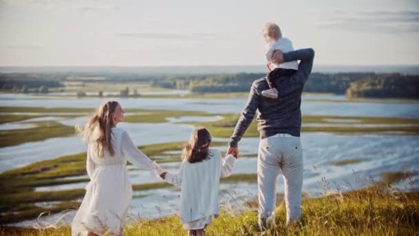 Ung familj står på fältet och håller hand medan du tittar på floden — Stockvideo