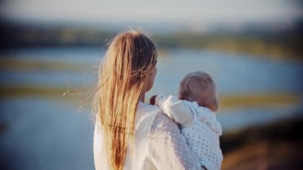 Młoda kobieta trzyma swoje dziecko stojąc na łonie natury i patrząc na rzekę — Wideo stockowe