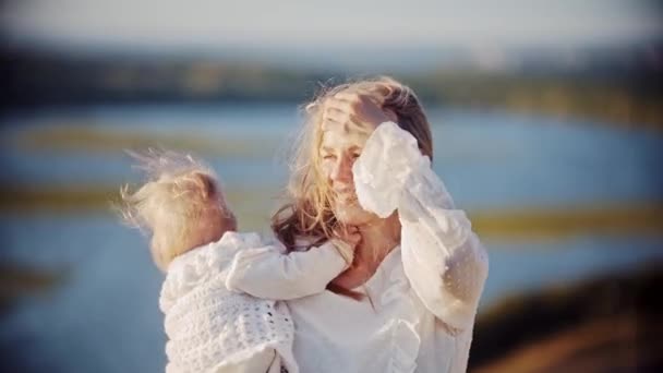 Mujer sonriente sosteniendo a su bebé mientras está de pie en la naturaleza y mirando al río — Vídeos de Stock