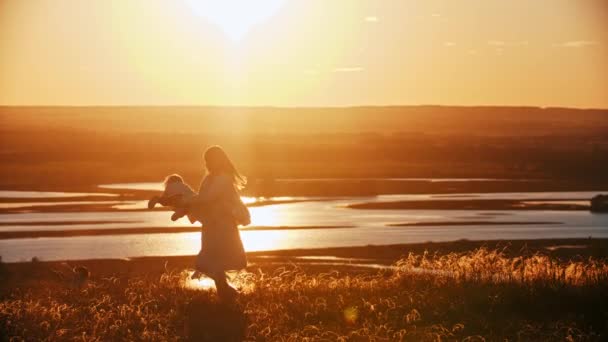 Jovem mulher girando seu bebê enquanto está de pé na natureza ao pôr do sol — Vídeo de Stock