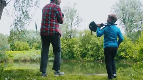 Pesca en la naturaleza: dos hermanos caminando hacia el río que sostienen instalaciones de pesca y un niño pequeño despliega una silla portátil — Vídeo de stock