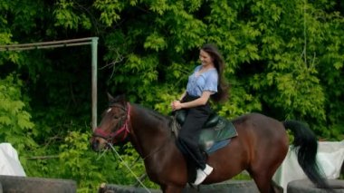 Genç, gülümseyen, uzun saçlı, mavi gömlekli mutlu kadın ata biniyor.