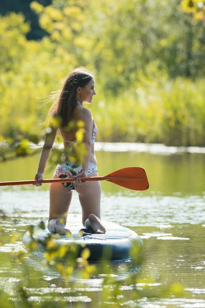 Молода жінка в квітковому купальнику стоїть на колінах на надувному човні і тримає весло за спиною — стокове фото