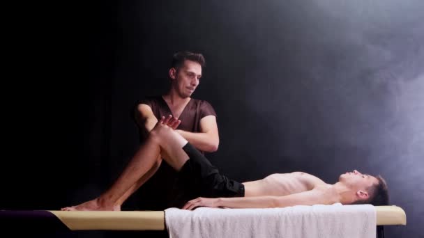 Sesja masażu w ciemnym studio - masażysta trzymający mans kolana i rozmawiający z nim — Wideo stockowe