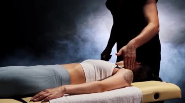 Karanlık stüdyoda masaj seansı. Terapist, müvekkiline krem sürüyor ve kadınların boynuna masaj yapıyor.