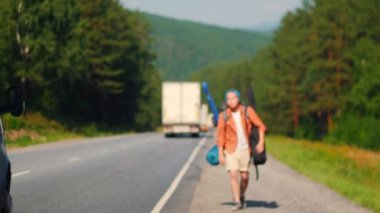 Büyük sırt çantalı sakallı bir adam yolda yürüyor ve bir araba yakalıyor.