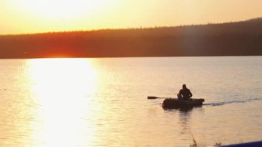 Baba ve oğul gün batımında göl kıyısında şişme bir teknede yelken açıyorlar.