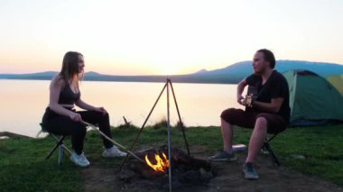 Genç adam ve kadın kampta ateşin başında oturuyor. Gitar çalan adam.