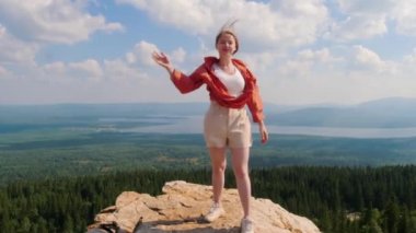 Dağın kenarında duran ve elini kameraya sallayan genç bir kadın.