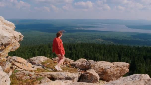Mujer joven de pie en el borde de la montaña y mirando al bosque — Vídeos de Stock