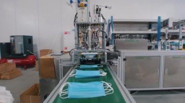 Mavi koruyucu tıbbi maskelerin endüstriyel üretimi. Koruyucu kostümlü ve eldivenli işçiler maskeleri bir araya topluyor.