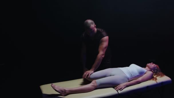 Mulher deitada em um sofá e um quiroprático trabalha com as pernas no estúdio escuro dobrando a perna no lado — Vídeo de Stock