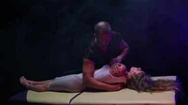 Karanlık stüdyoda kayropraktik terapi. Genç bir kadını belli bir pozisyona sokup omzuna baskı yapan bir erkek terapist.
