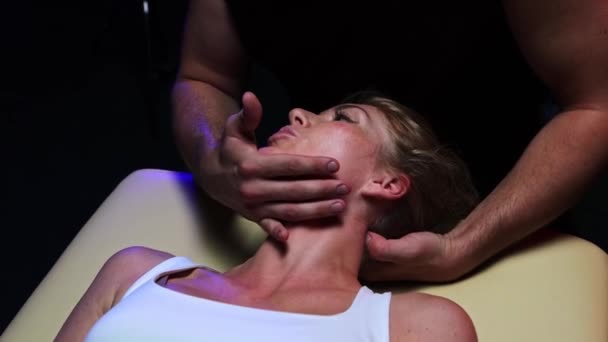Хиропрактика в темной студии - мужчина-терапевт расслабляет шею молодой женщины и поворачивает голову в сторону — стоковое видео