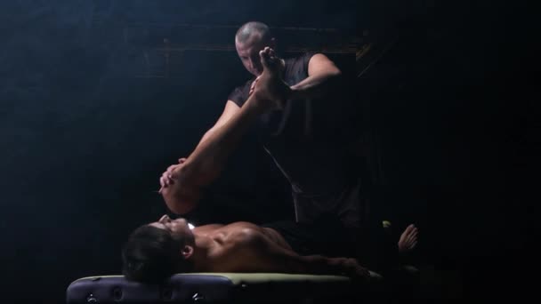 Chiropractische therapie in de donkere studio - een therapeut die op het been van zijn cliënt drukt naar zijn lichaam — Stockvideo