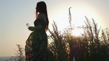 Günbatımında elinde çiçekle suyun kenarında duran gevşek elbiseli genç romantik kadın.