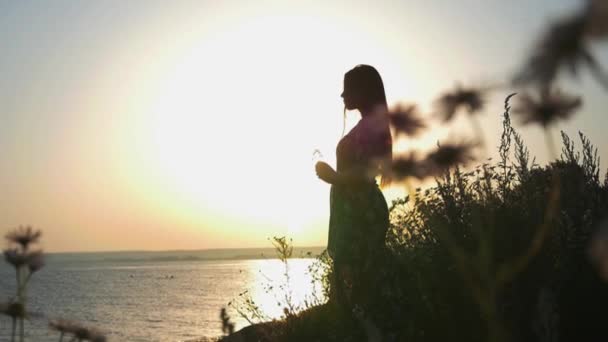 Mujer romántica joven en vestido suelto de pie junto al agua en la puesta del sol — Vídeo de stock
