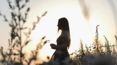 Gün batımında elinde çiçekle tarlada yürüyen bol elbiseli genç bir kadın.