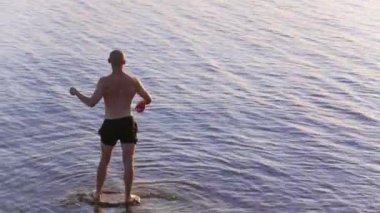 Genç, kel, beyaz adam nehirde duruyor ve suya taş atıyor.