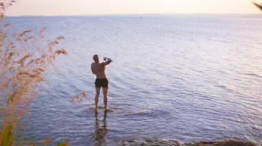 Genç kel adam nehirde duruyor ve suya taş atıyor.