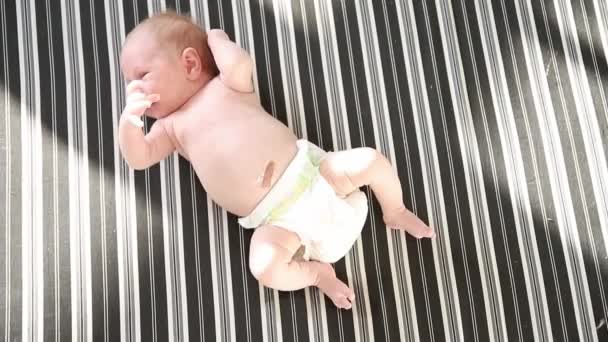 Aranyos aktív újszülött egy fehér pelenkában fekszik az ágyon csíkos lepedőn és mozog — Stock videók