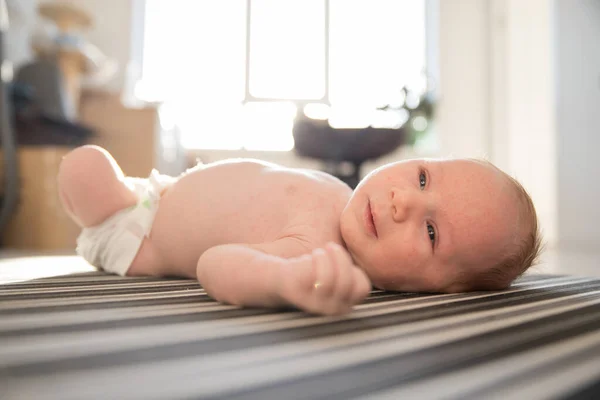 Um pequeno bebê bonito em uma fralda inquieto deitado de costas em lençóis listrados à luz do dia — Fotografia de Stock