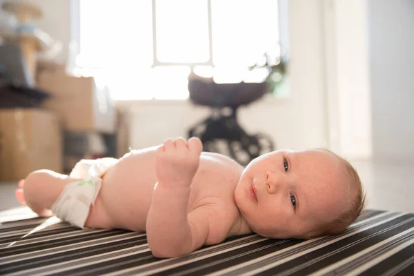 En liten söt bebis ligger rastlöst på rygg på randiga lakan i dagsljus — Stockfoto