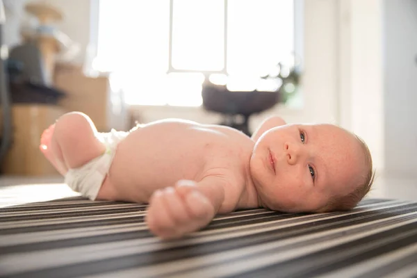 白天，一个穿着尿布的小宝宝躺在带条纹的床单上，不安分地躺在床上 — 图库照片