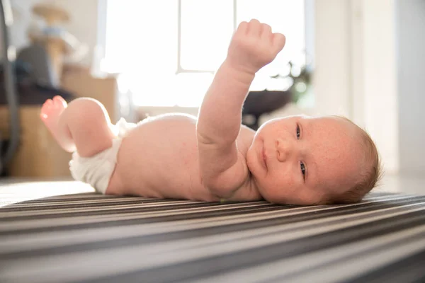 Een pasgeboren schattige baby in een luier rusteloos liggend op zijn rug op gestreepte lakens in daglicht — Stockfoto