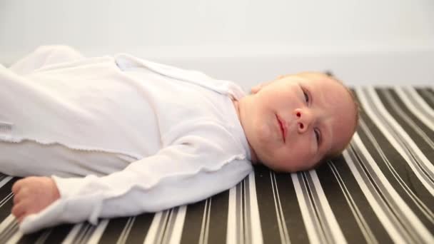 Un petit nouveau-né en costume blanc couché sur le dos sur un drap rayé sur un lit — Video