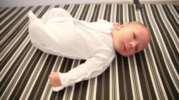 Een pasgeboren baby in wit kostuum liggend op zijn rug op gestreept laken op een bed — Stockvideo