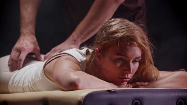Массаж в темной современной студии - женщина лежит на диване, а терапевт работает с ее телом - массирует поясницу — стоковое видео