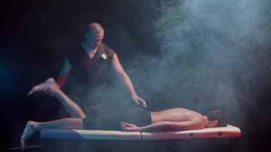 Karanlık stüdyoda kiropraktik terapi. Bir terapist müvekkilini kanepeye oturtup bacaklarını oynatıyor.