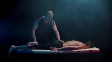 Karanlık stüdyoda kiropraktik terapi. Bir terapist müvekkilini kanepeye oturtup bacaklarını ısıtıyor.